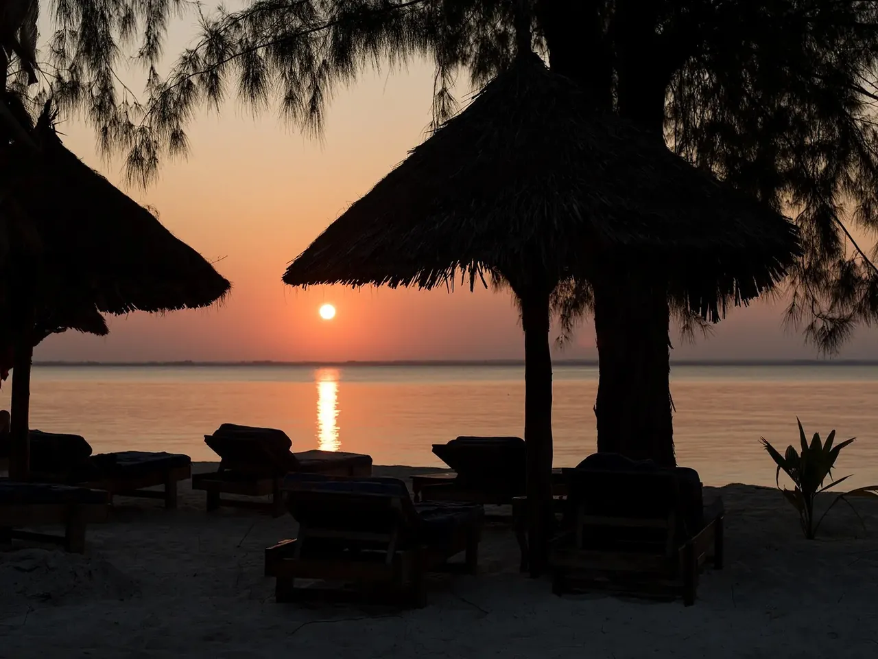 Thatched beach umbrellas and loungers silhouetted against an orange sunset over the bay.