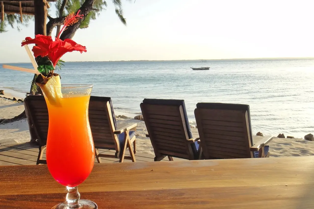 An orange and red cocktail with a hibiscus flower on a bar counter, with deckchairs and calm sea behind.