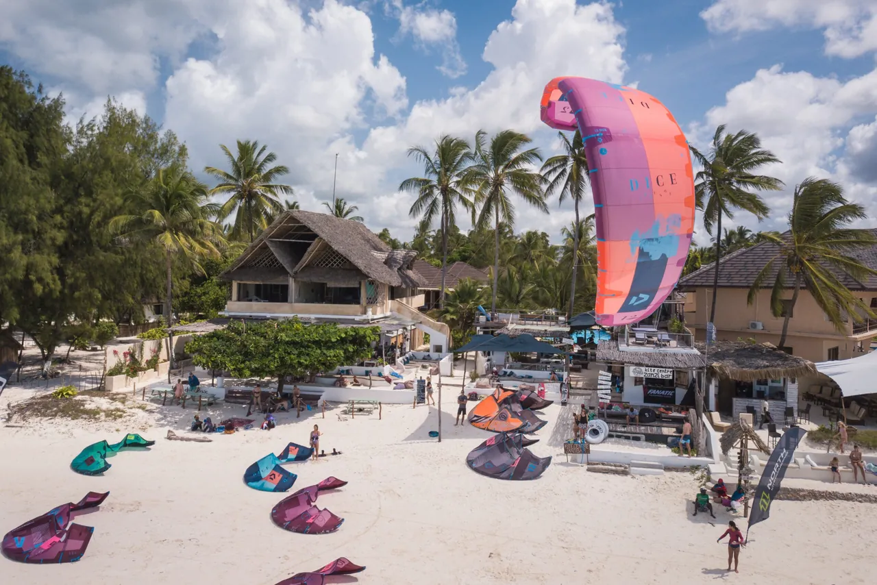 Kites laid out on a white beach in front of thatched beachfront buildings and palms.