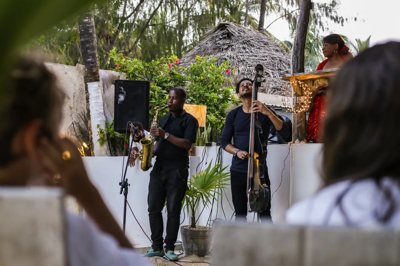 A band with saxophone and double bass playing outdoors in front of a thatched building, seen past audience members.