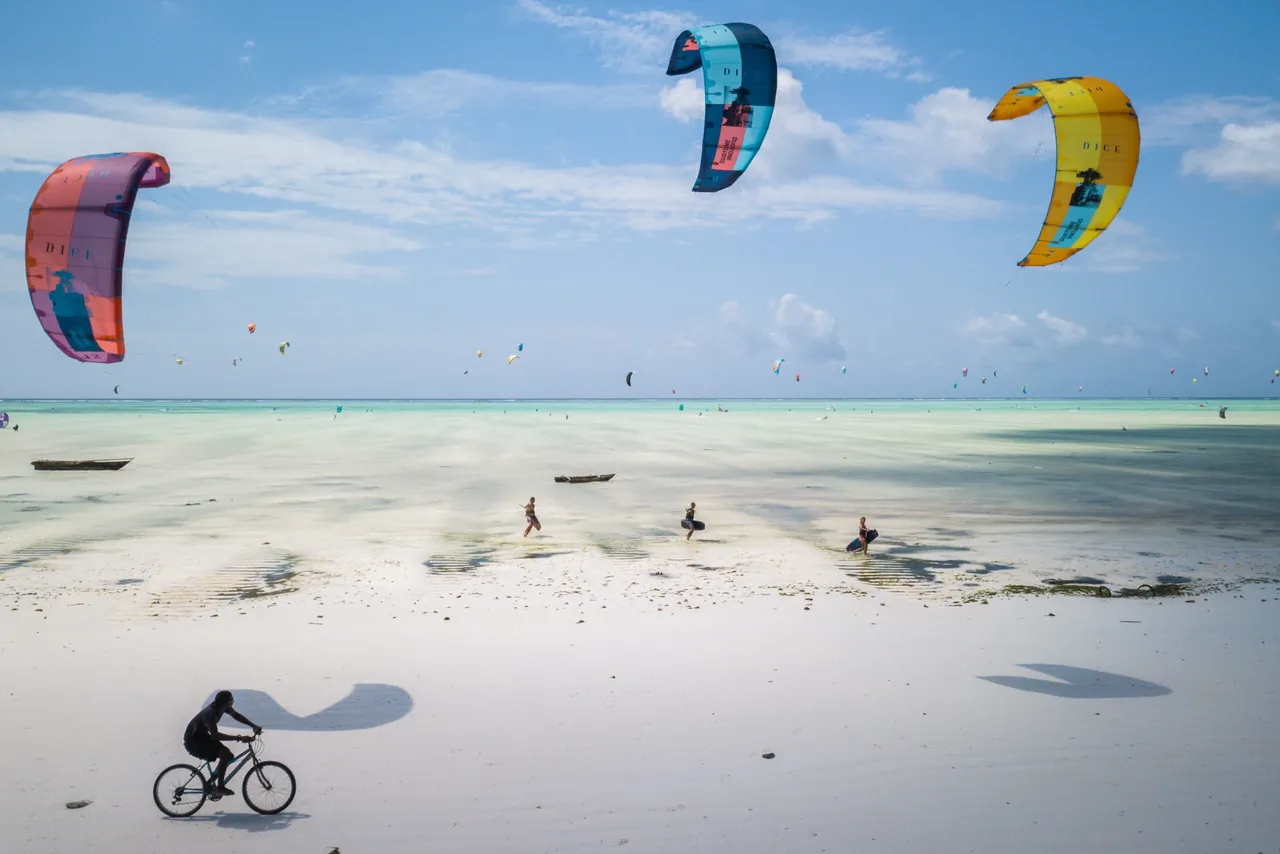 Colourful kites in the sky above a wide white beach at low tide, with a cyclist in the foreground.