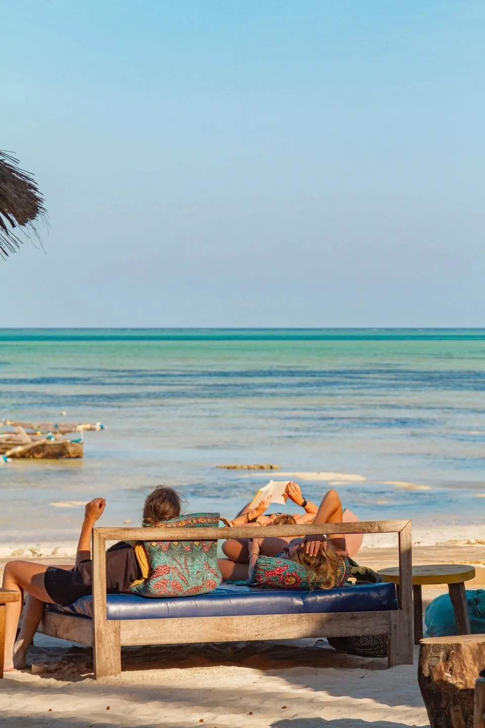 Two people relaxing on a cushioned wooden daybed on the sand, the turquoise sea and moored boats beyond.
