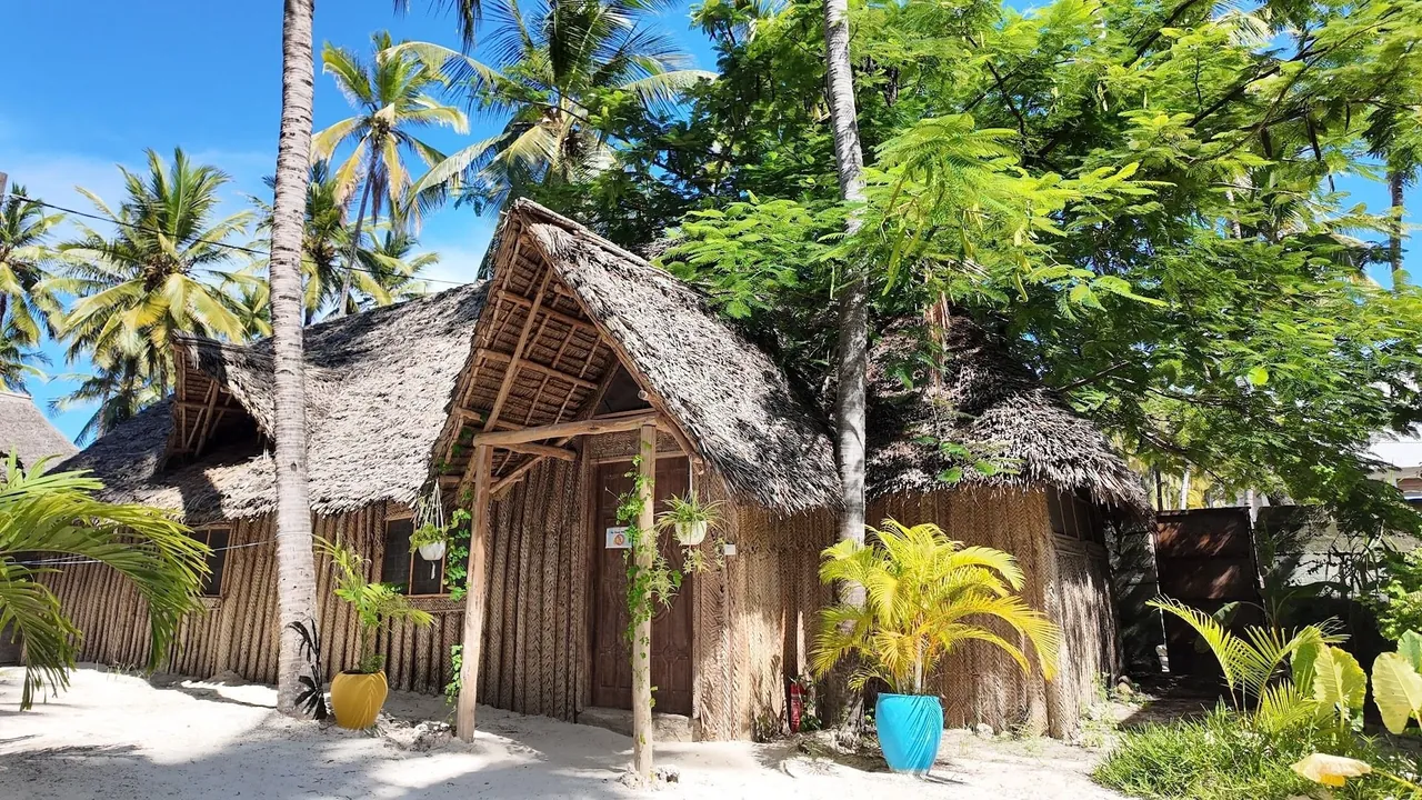Thatched bungalows among palms and tropical plants on sand.