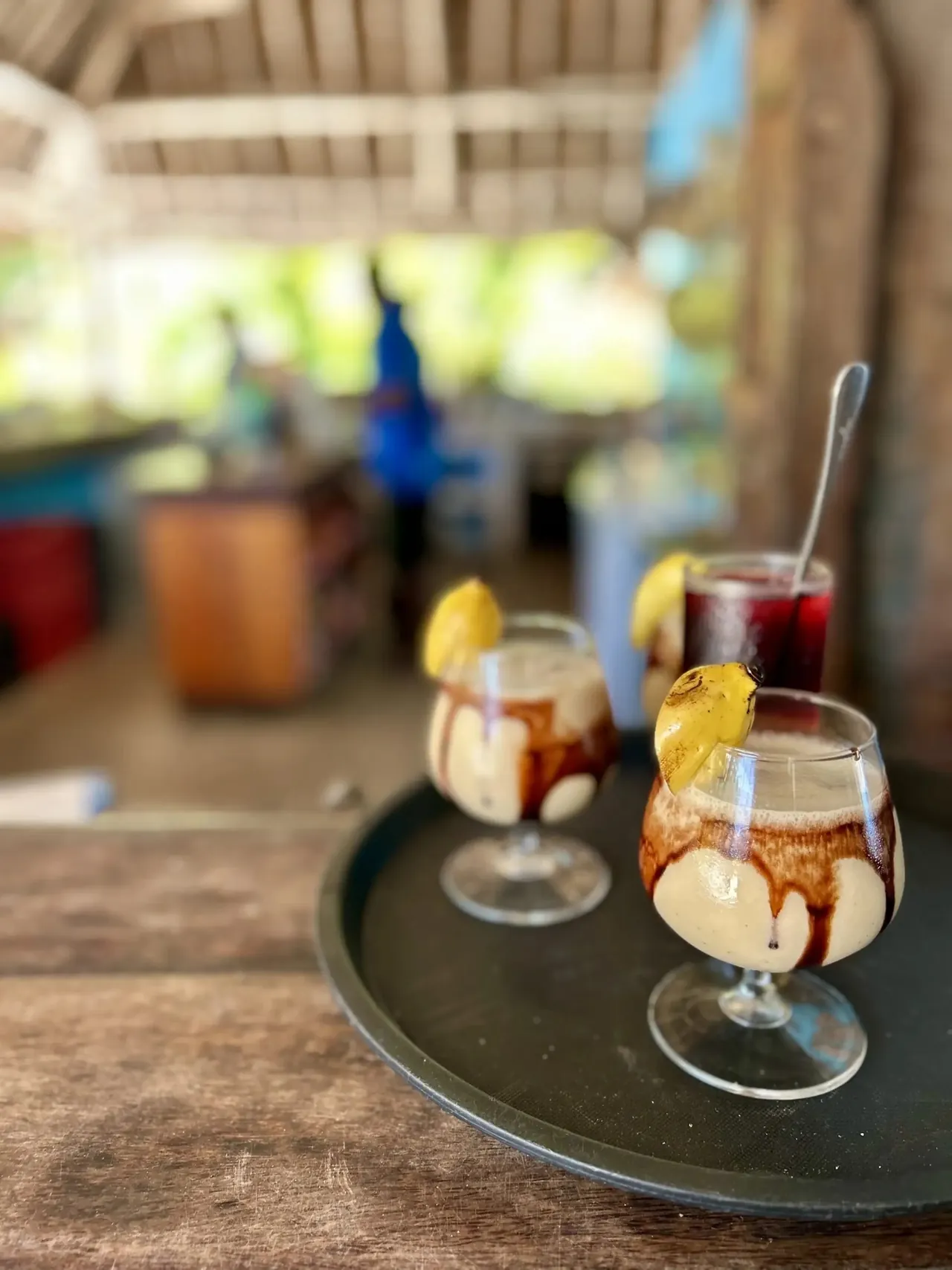 A tray with two iced coffee or cocktail glasses on a bar, a blurred thatched bar behind.
