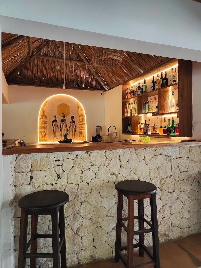 A bar counter with a stone front, two tall stools and shelves of bottles under a thatched roof.