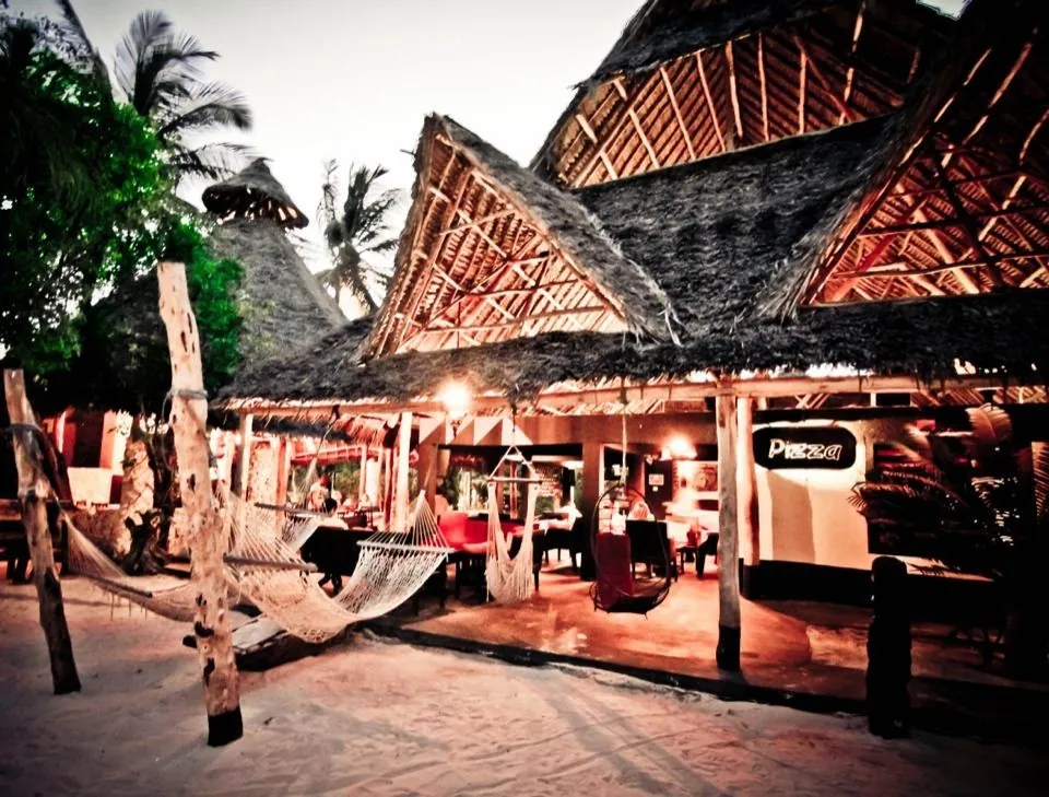 Thatched buildings at dusk with a hammock in the foreground and warm lights inside the bar.