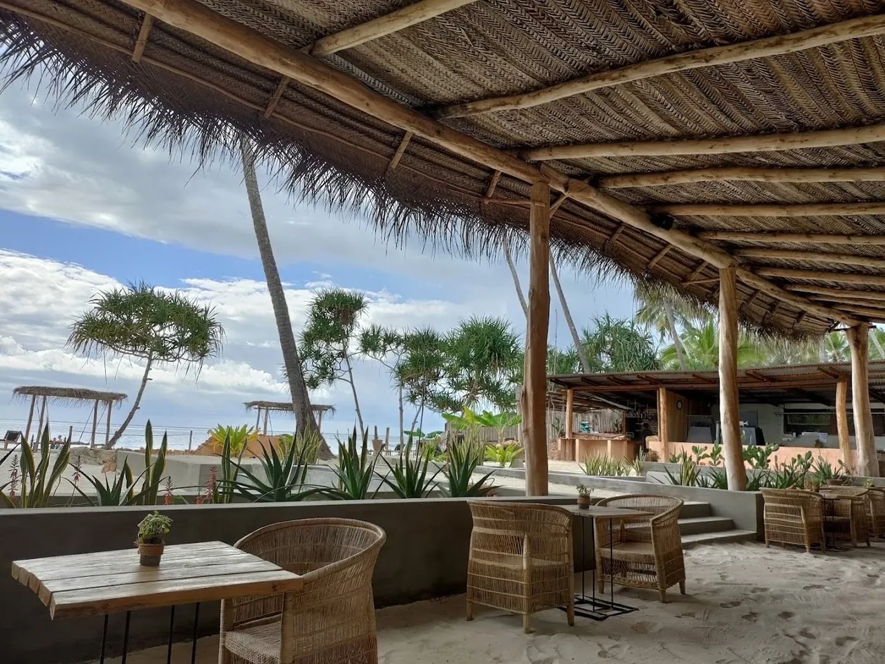 An open-sided thatched lounge with rattan chairs and wooden tables, looking out to palms and the beach.
