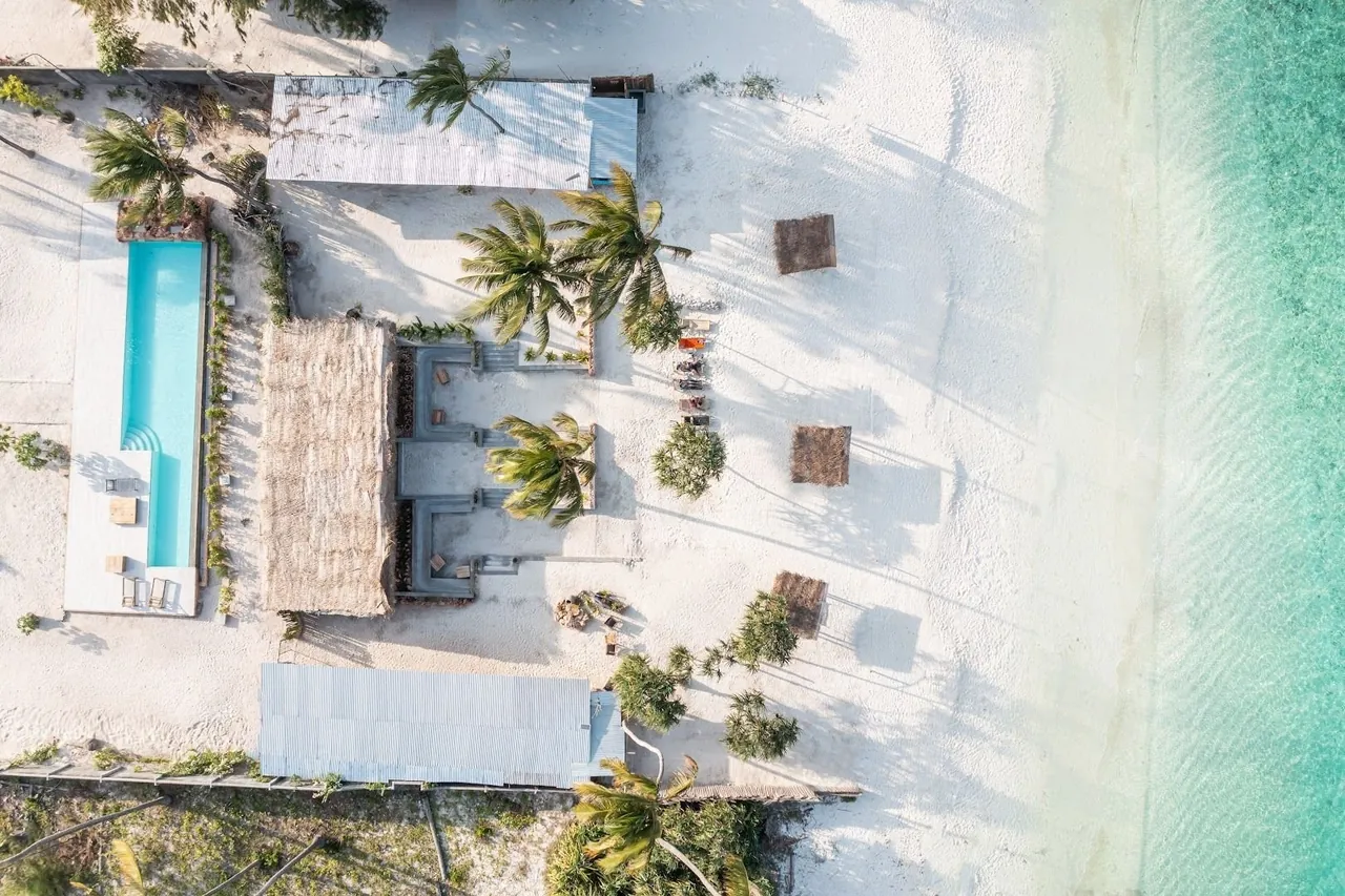 Aerial view of a thatched beach lounge, wooden cabanas and a pool beside a white beach and turquoise shallows.