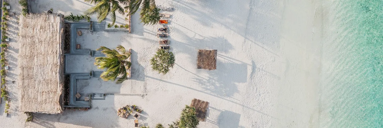 Aerial panorama of white sand with a thatched pavilion, cabanas and palm trees beside turquoise water.