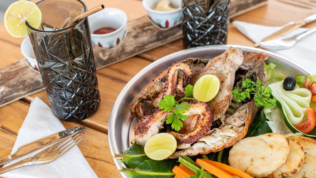 A grilled seafood platter with octopus, fish and chapati on a wooden table, with a cocktail glass alongside.