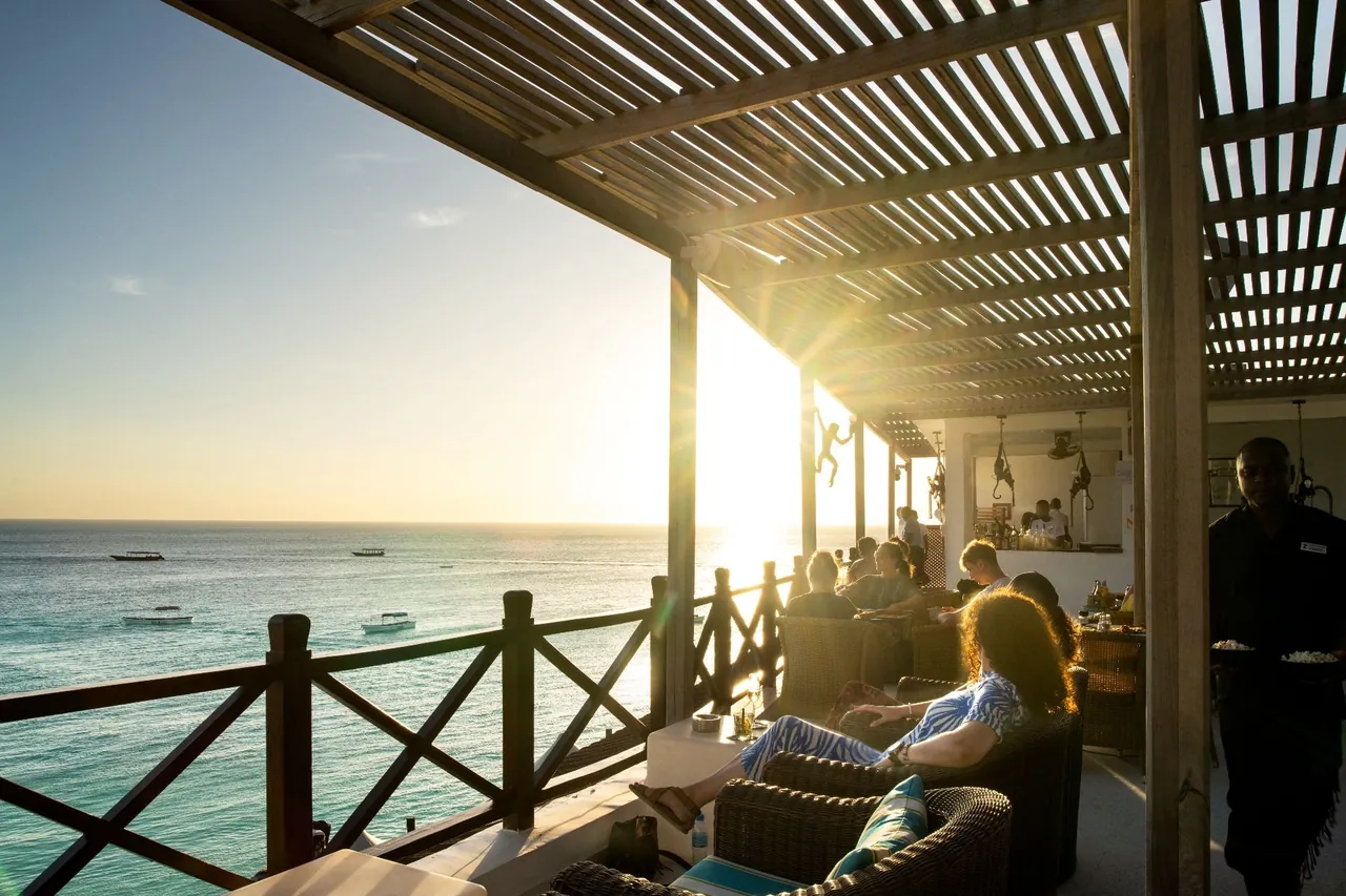 A covered rooftop terrace with guests on sofas as the sun sets over the ocean