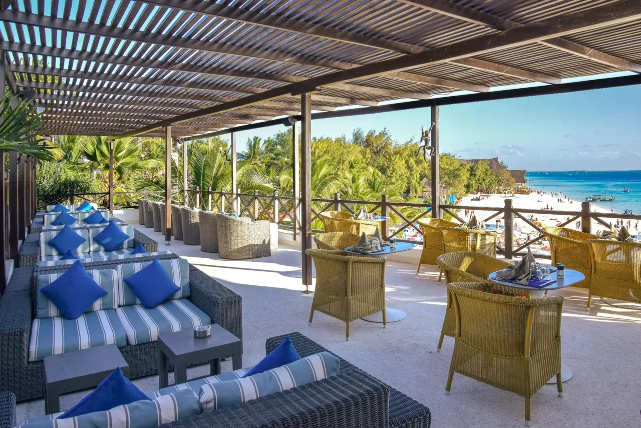 A shaded terrace with blue-and-white striped sofas and wicker chairs facing the beach