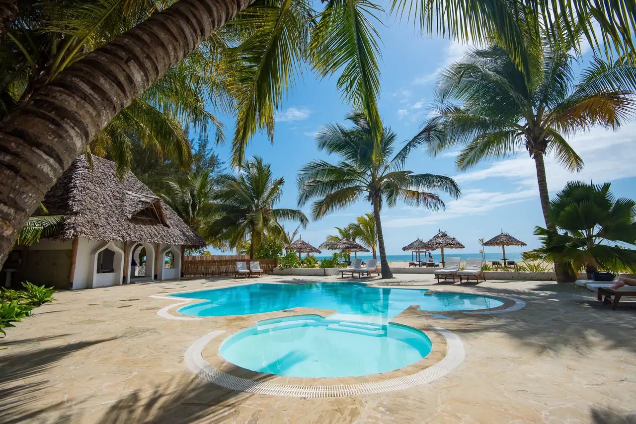 The hotel pool under palm trees, with thatched umbrellas on Paje beach beyond