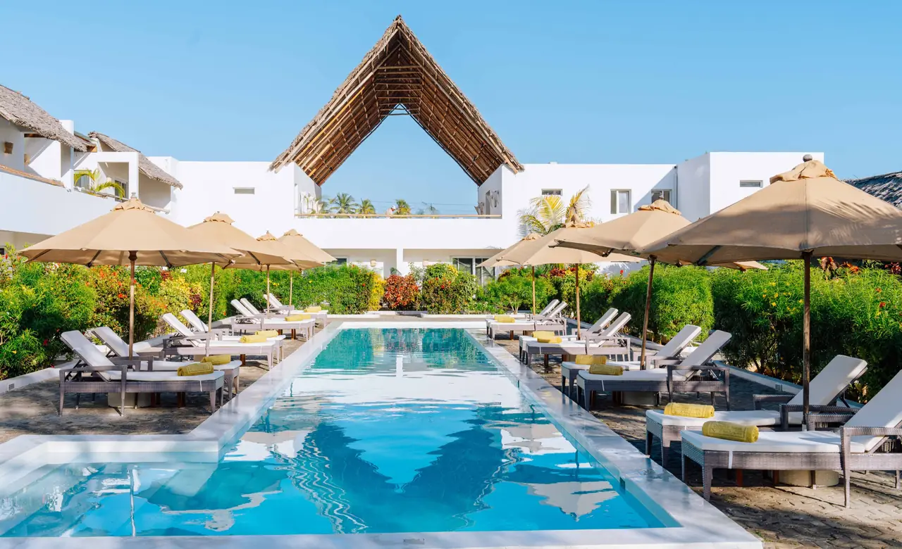 A long pool lined with loungers and umbrellas, leading to an A-frame thatched building