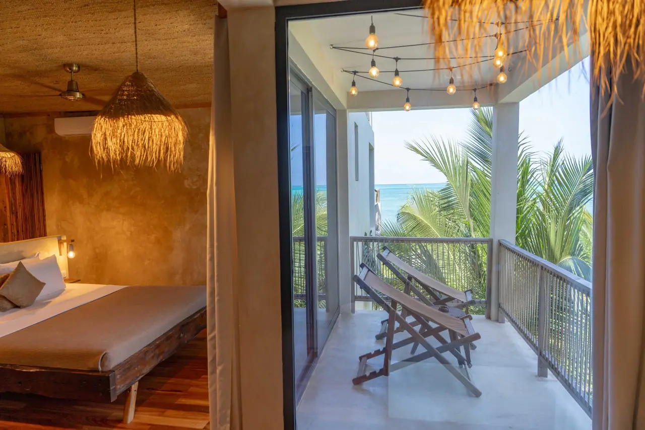 A guest room opening onto a balcony with a lounger and sea views through the palms