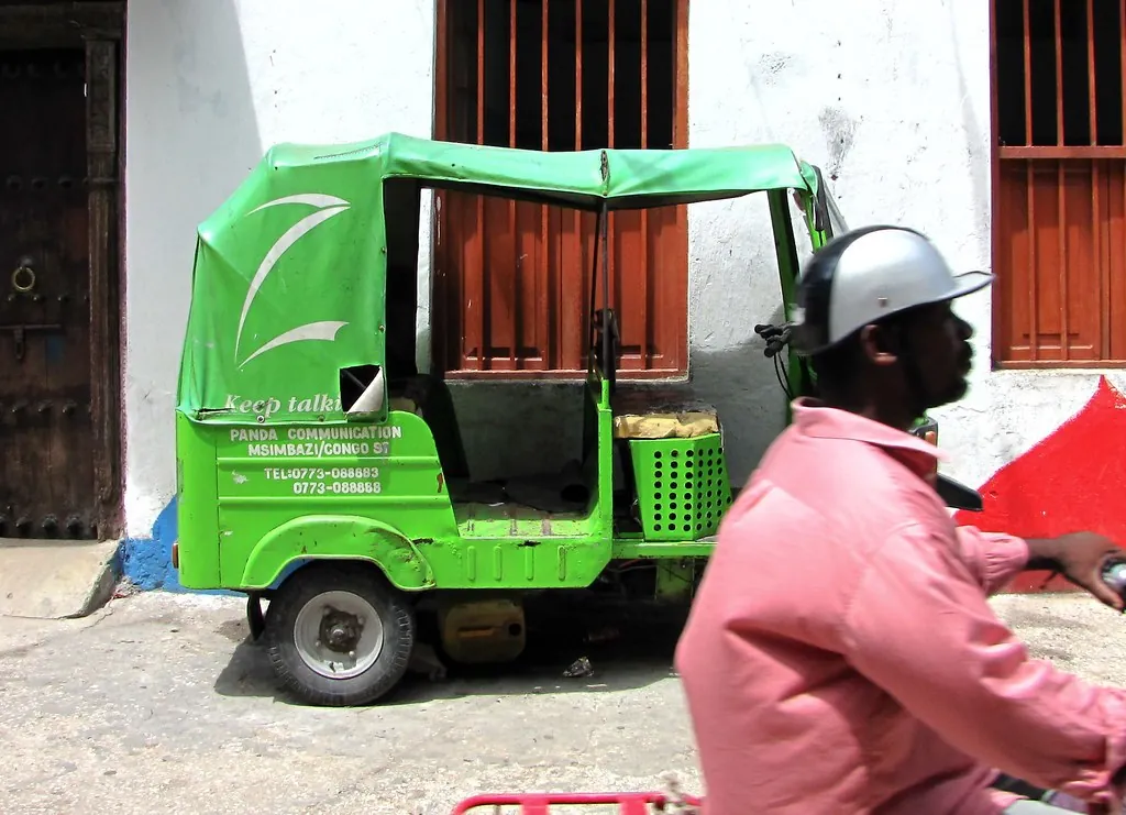 Green advertising-livery bajaji parked against a white wall, with a motorcyclist passing