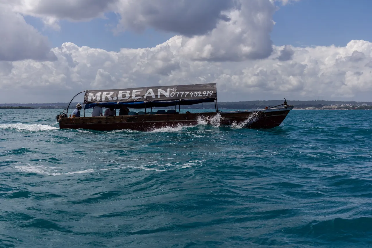 Wooden motor boat with a painted canopy and a few passengers crossing choppy water, with the Zanzibar shore behind