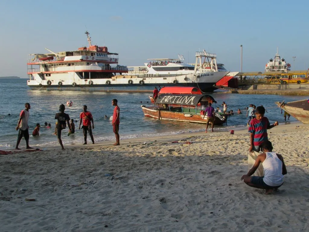 Stone Town beach in late afternoon: a canopy boat in the shallows, young men playing football, and passenger ferries at the terminal behind