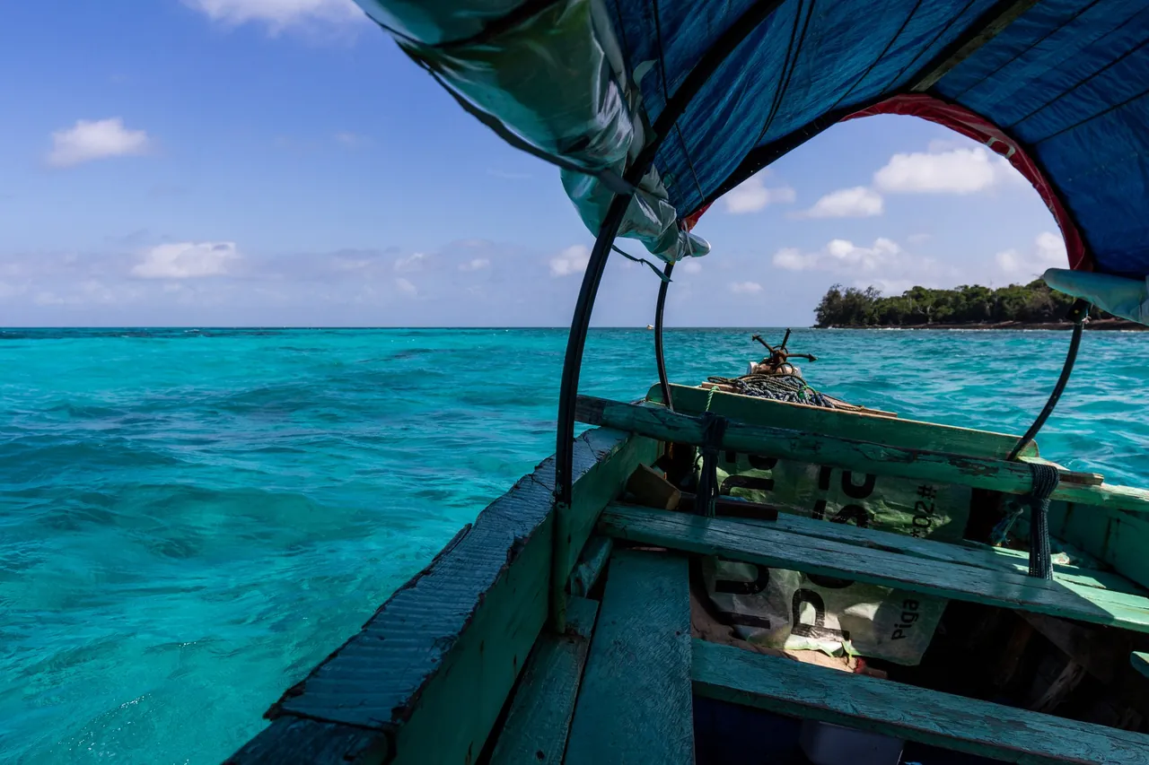 View from inside a wooden canopy boat with green-painted benches, approaching a small wooded island across turquoise water