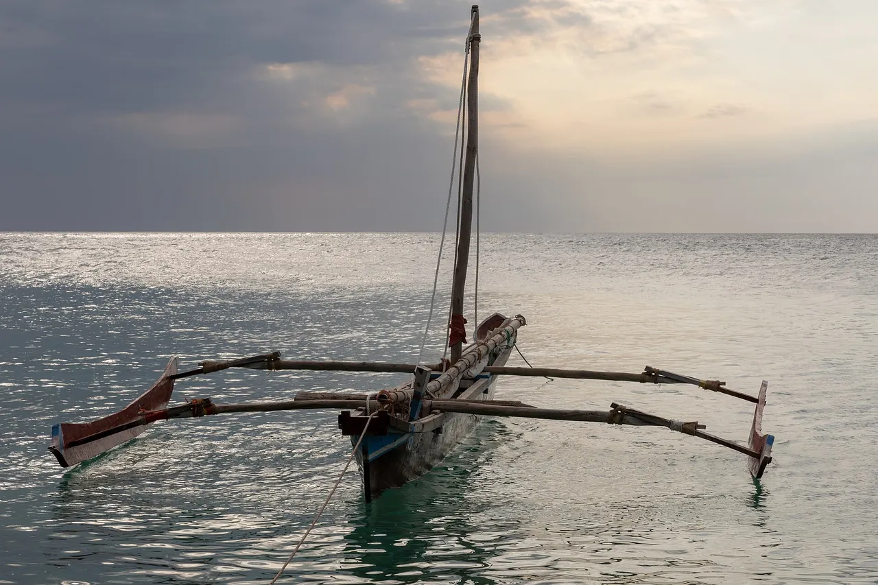 Traditional wooden outrigger sailing canoe (ngalawa) moored on silvery water under a cloudy sky