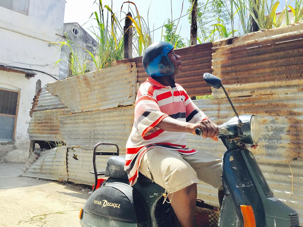 Man wearing a blue helmet riding a scooter past corrugated-iron fencing