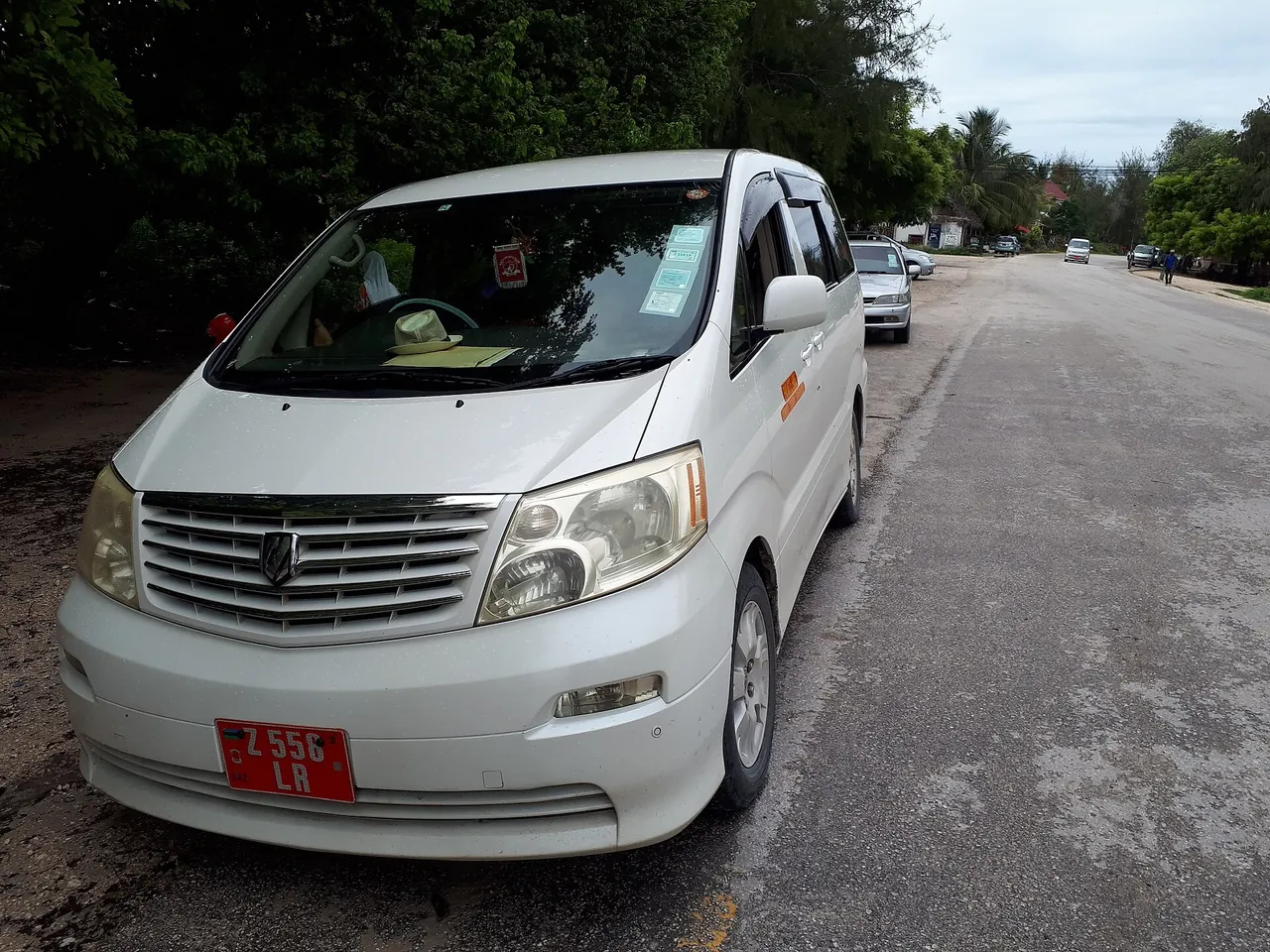 A white Toyota Alphard minivan with a red Zanzibar number plate parked at the edge of a tarmac road in Paje