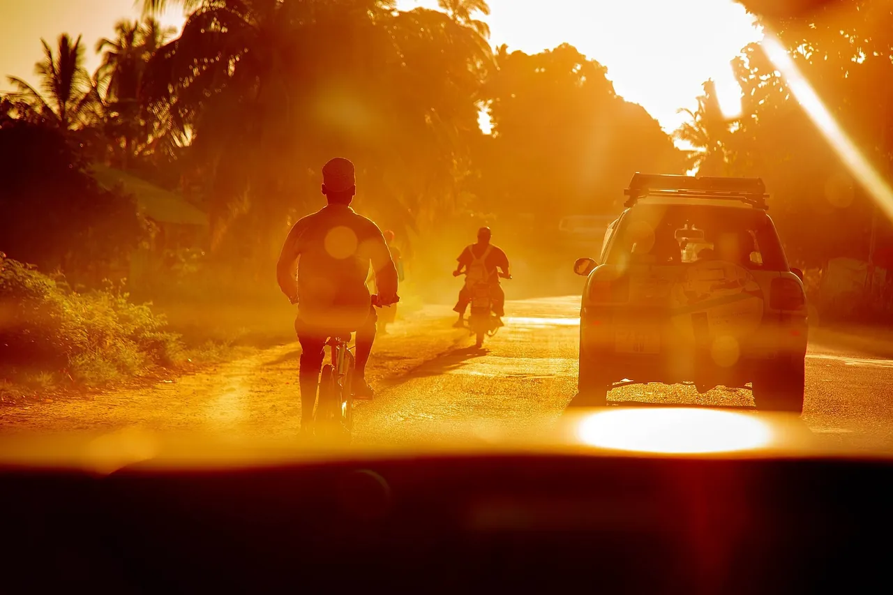 View through a car windscreen at sunset of a cyclist, a motorcyclist and an SUV sharing a road in Fuoni