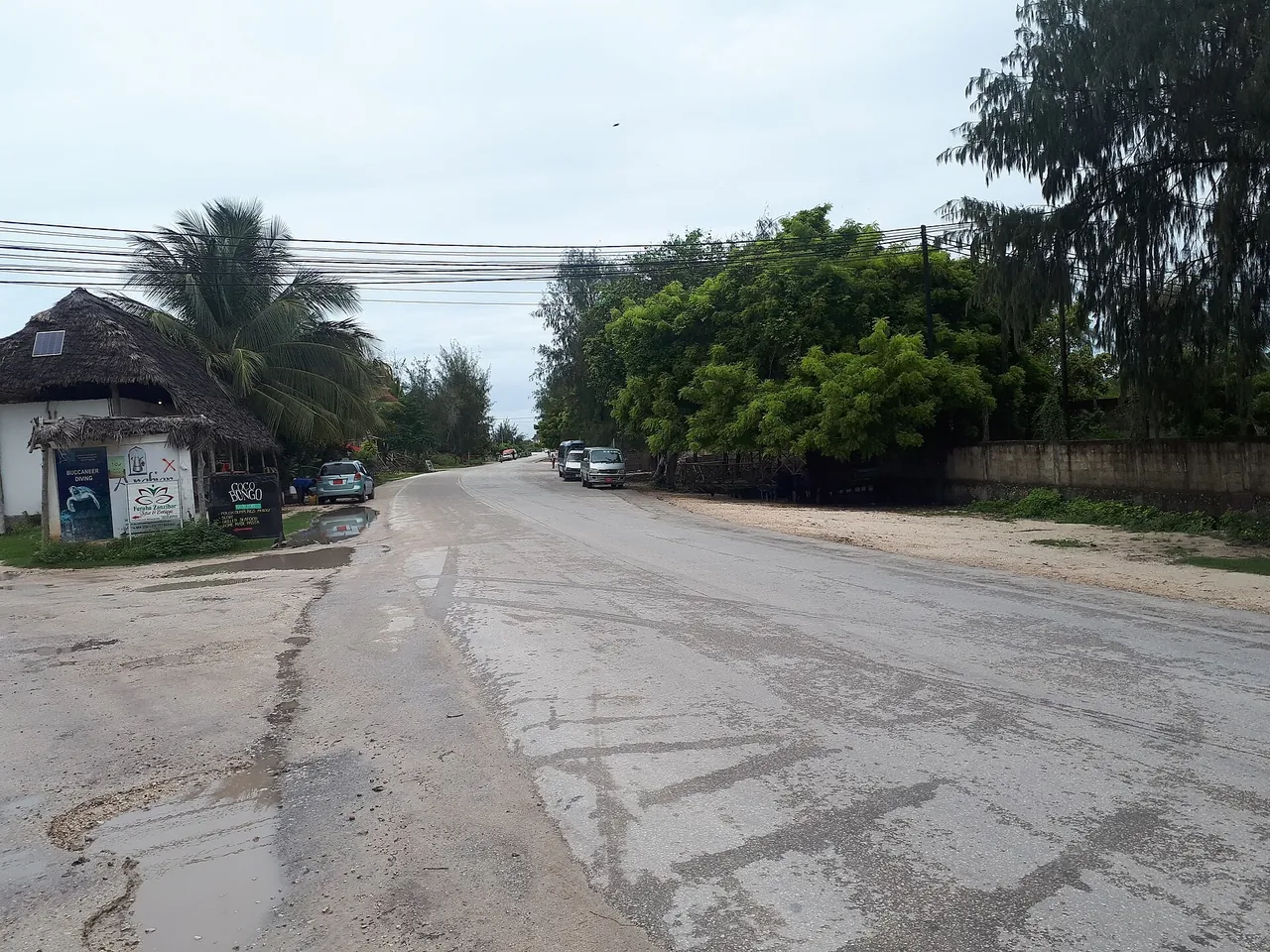 A wet, patched tarmac road through Paje with puddles at the verge, parked minibuses and a thatched building