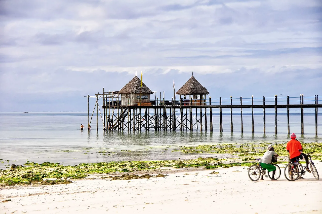 Two cyclists paused on a white beach at low tide, looking towards a long wooden pier with two thatched huts