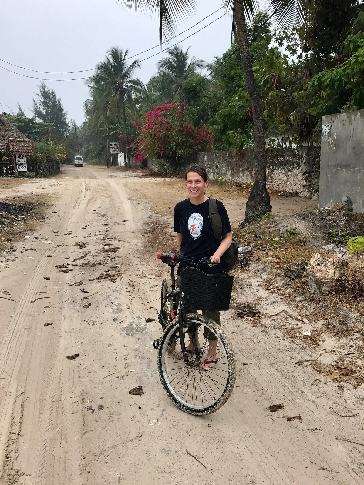 A woman standing with a basket-fronted bicycle on a sandy village lane lined with palms, with lodge signs behind