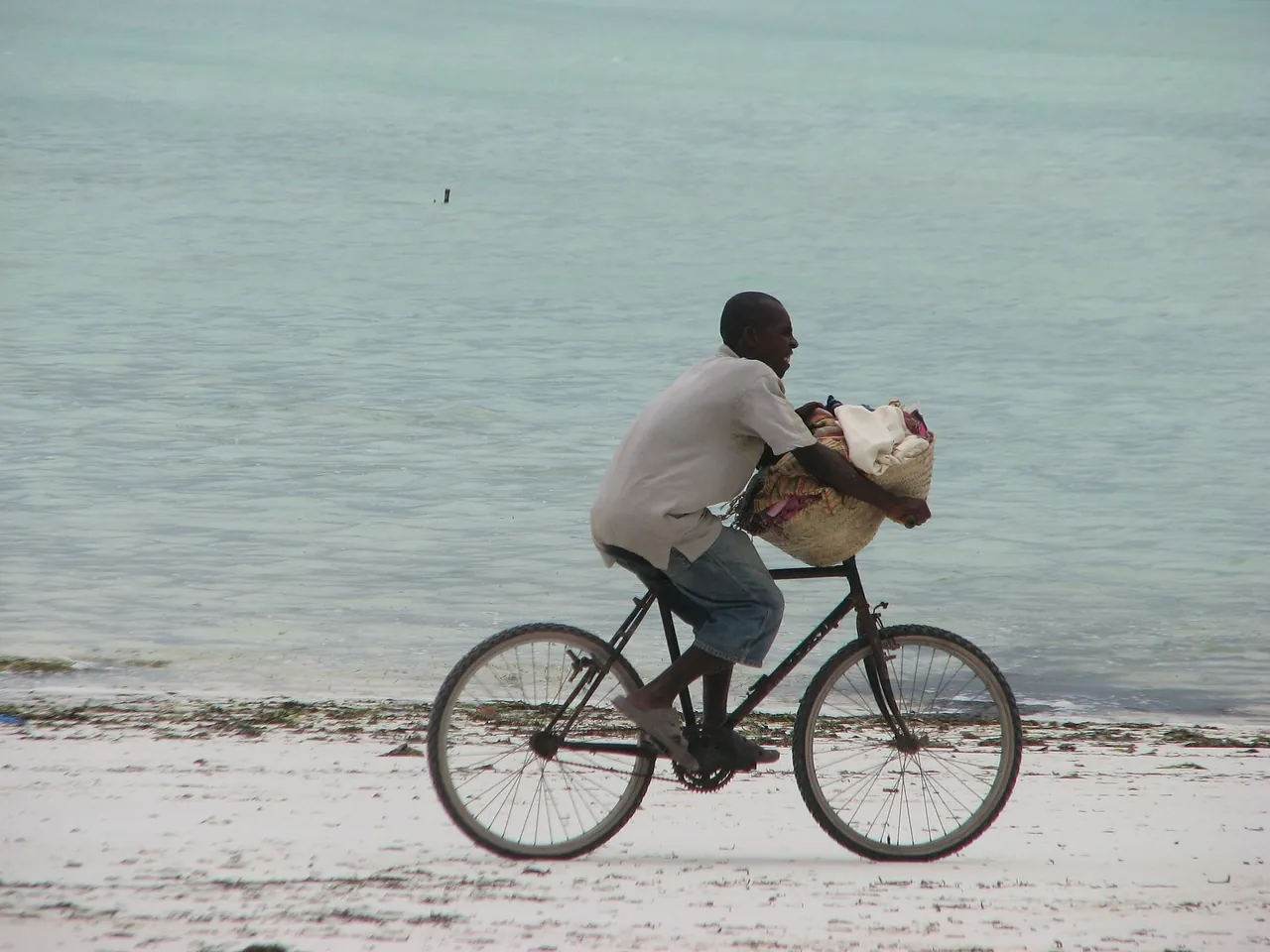 A young man cycling along a pale beach carrying a large woven basket of fabrics