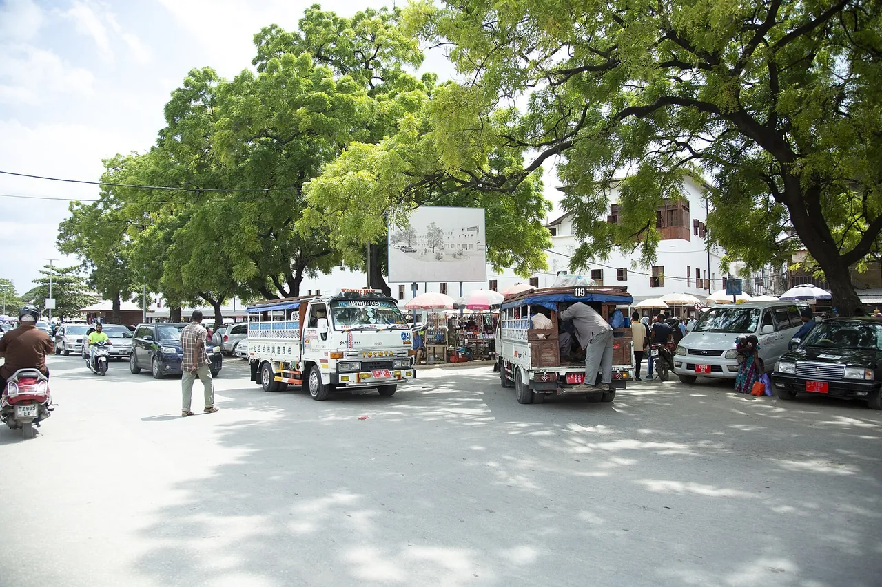 Tree-lined street with truck-bodied dala dalas, cars and a scooter