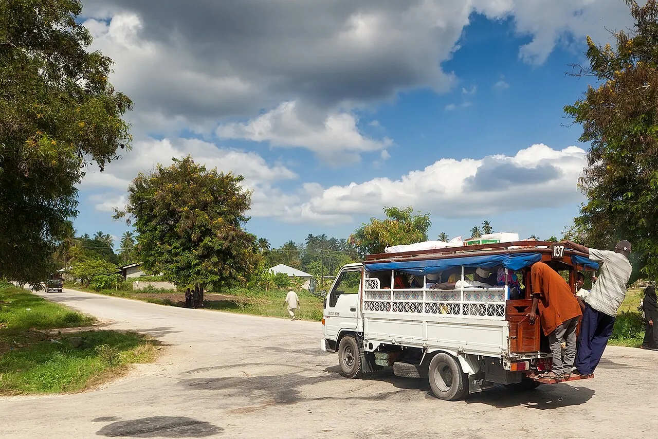 Small truck dala dala with passengers climbing aboard at the back, on a rural road