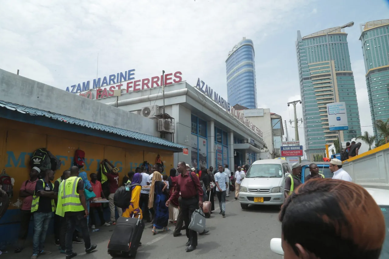 Crowded street outside the Azam Marine Kilimanjaro Fast Ferries terminal in Dar es Salaam, with men in numbered hi-vis vests and passengers wheeling suitcases