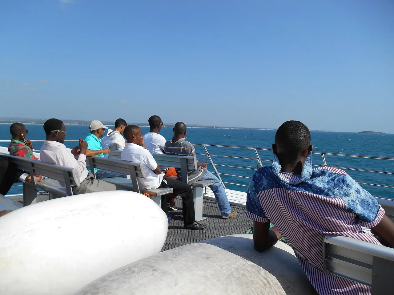 Passengers on benches on the open top deck of a fast ferry, looking out over blue sea towards a low coastline