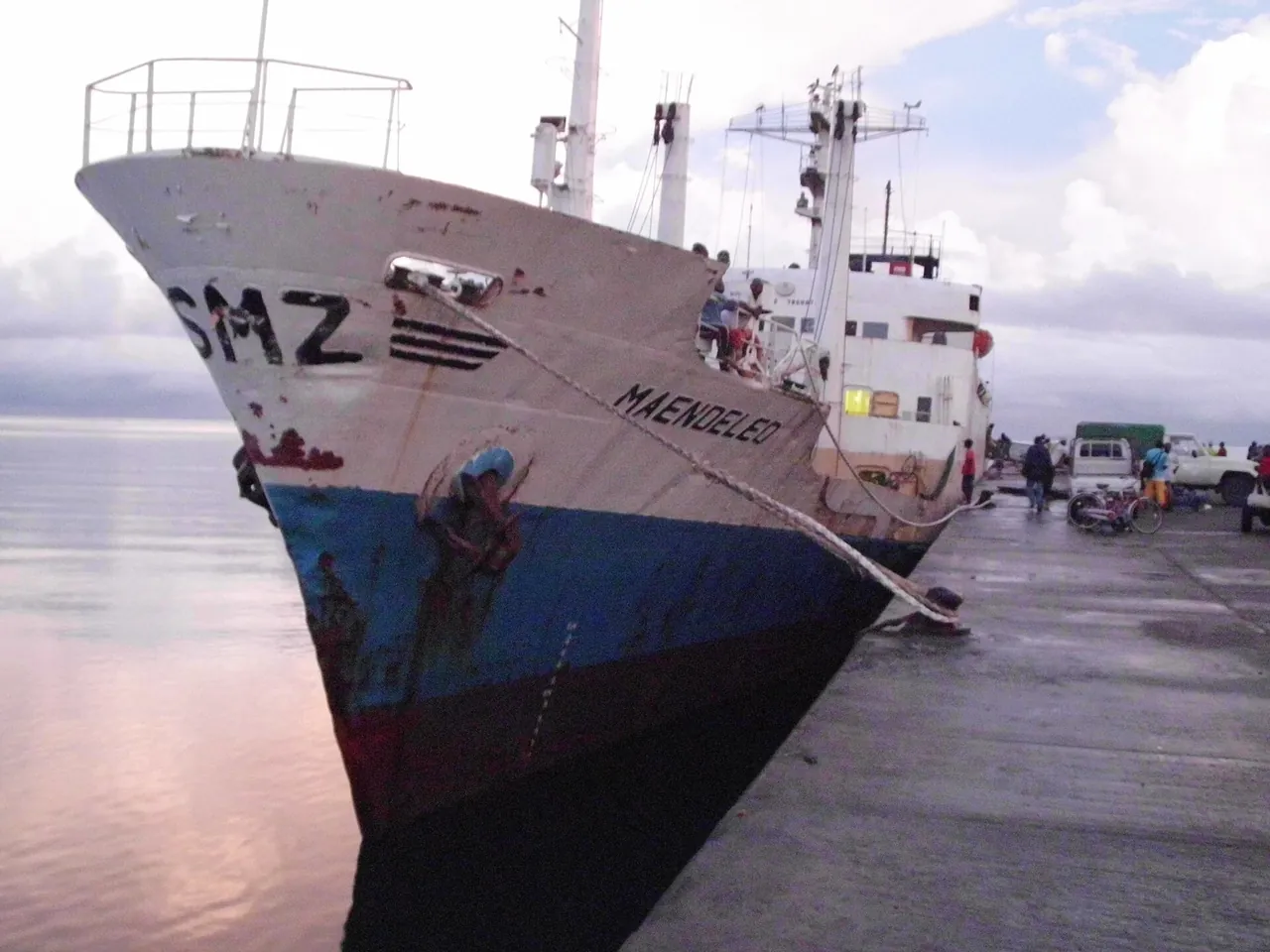 Bow of the weathered white-and-blue ship MV Maendeleo, marked SMZ, moored at a concrete wharf at dusk