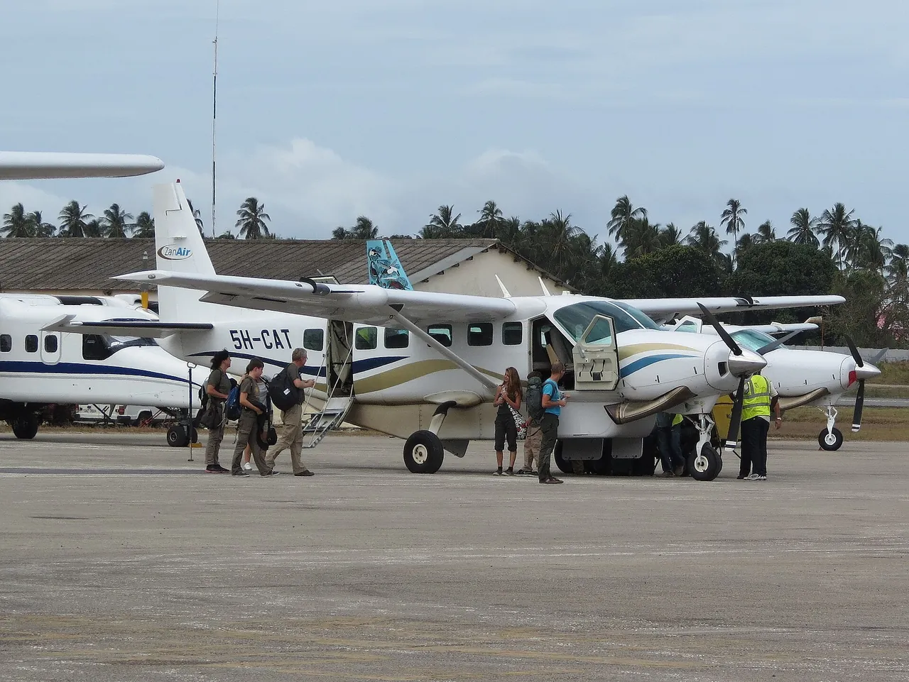 Passengers with backpacks boarding a ZanAir Cessna 208 Caravan, 5H-CAT, on the apron at Zanzibar airport, with palm trees behind