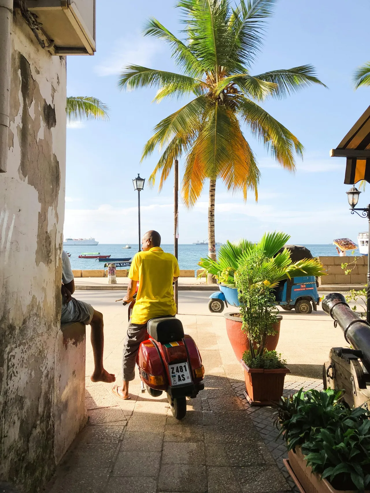 Man on a red scooter on the Stone Town seafront, with a blue bajaji marked taxi on the road beyond