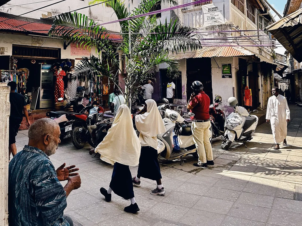 Busy Stone Town square with pedestrians and a row of parked motorbikes and scooters