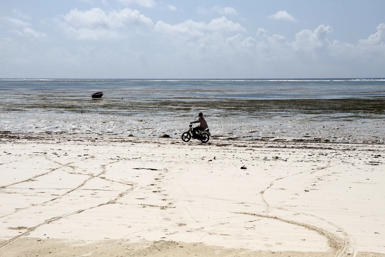 A man riding a moped along the edge of a wide white beach at low tide, with exposed seagrass flats behind