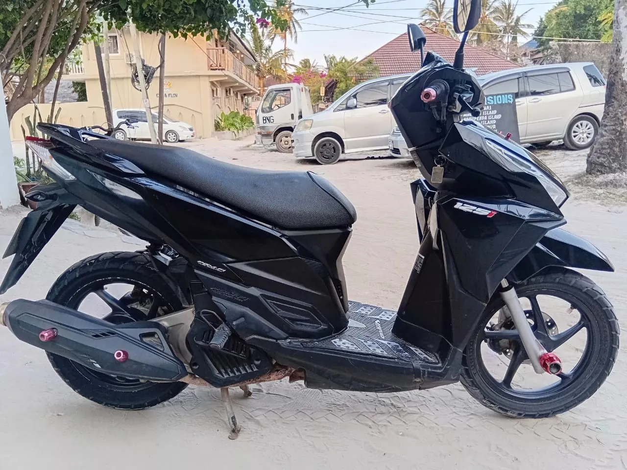 A black Honda Click 125i scooter parked on a sandy street with parked cars and a tour-excursions sign behind
