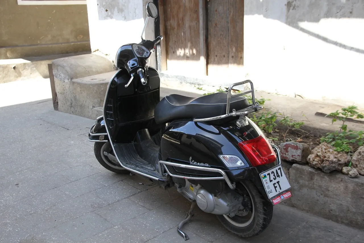 A black Vespa scooter with a Zanzibar 'Z 347 FT' number plate parked by a coral-stone bench and an old wooden door