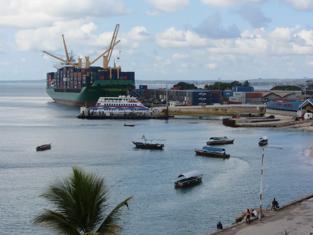 Elevated view of Zanzibar port: a container ship, a passenger ferry at a floating pontoon linked by a yellow walkway, and a blue terminal roof with 'Karibu Zanzibar' sign