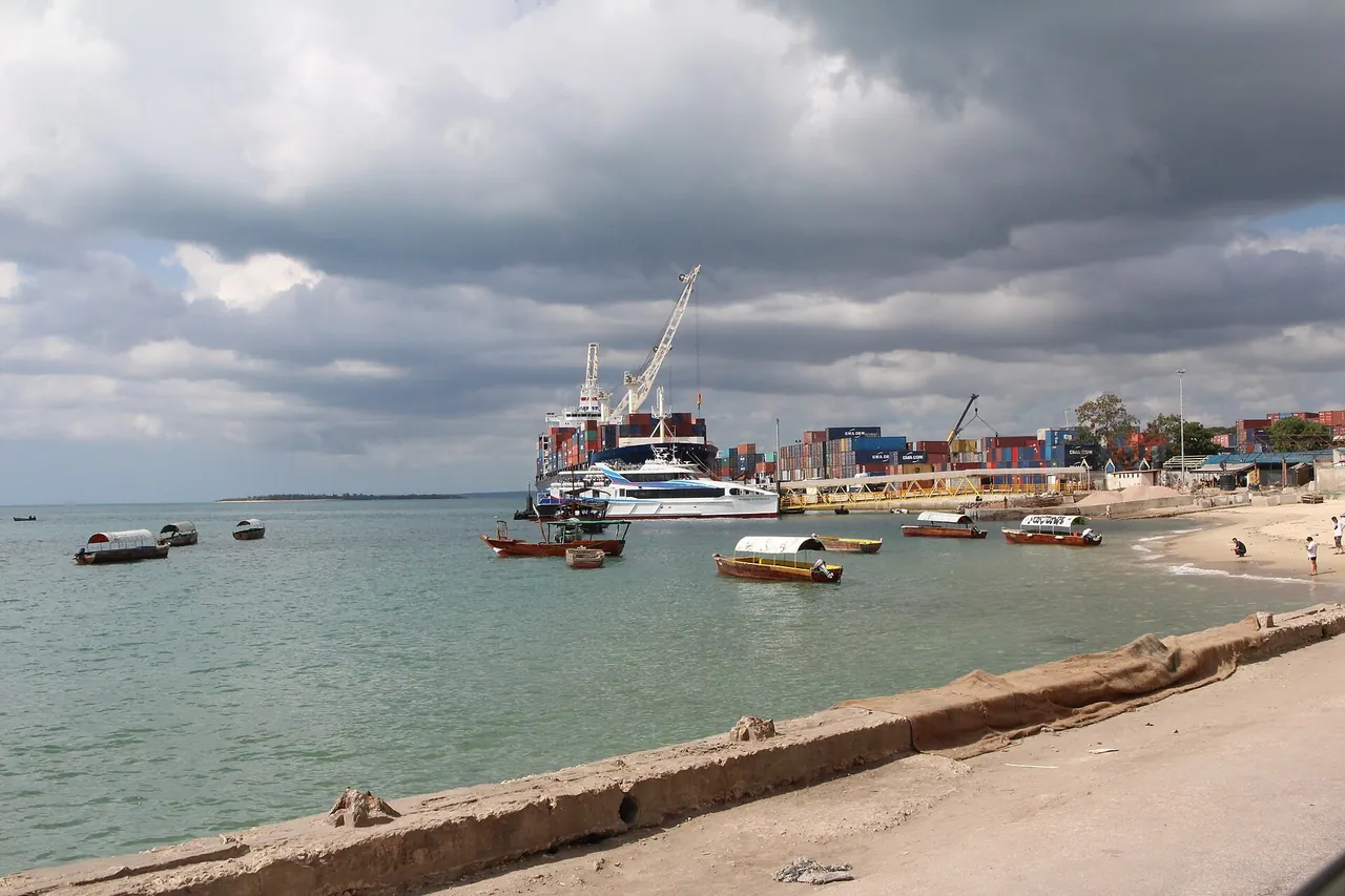 Passenger catamaran docked at the Malindi ferry terminal next to a container ship and stacked containers, with small canopy boats in the foreground