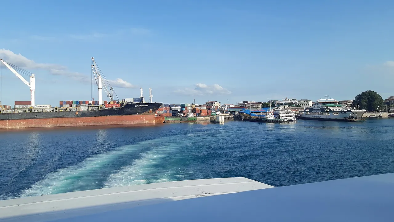 View from the stern of a departing ferry: wake in the foreground, Zanzibar port with a cargo ship, cranes and docked ferries behind