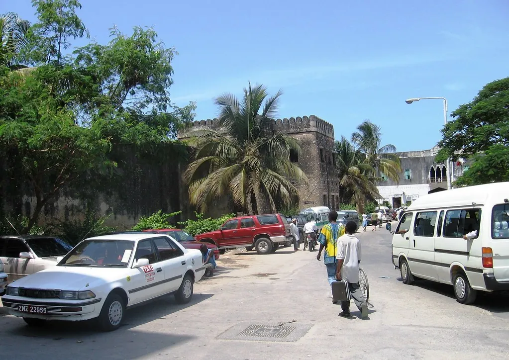 White saloon taxi with a roof sign parked on a Stone Town street, with an old fort wall behind