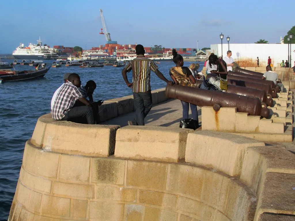 People sitting and strolling on a stone seawall lined with old cannon, with boats and the port behind