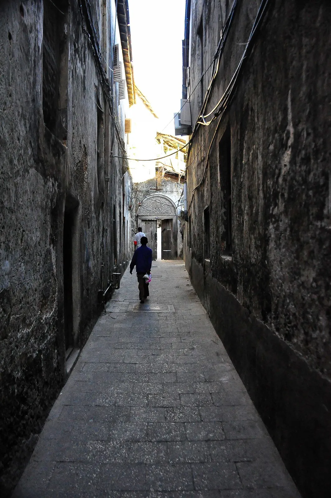 A narrow paved alley between tall weathered coral-stone walls, with people walking towards a carved arched doorway