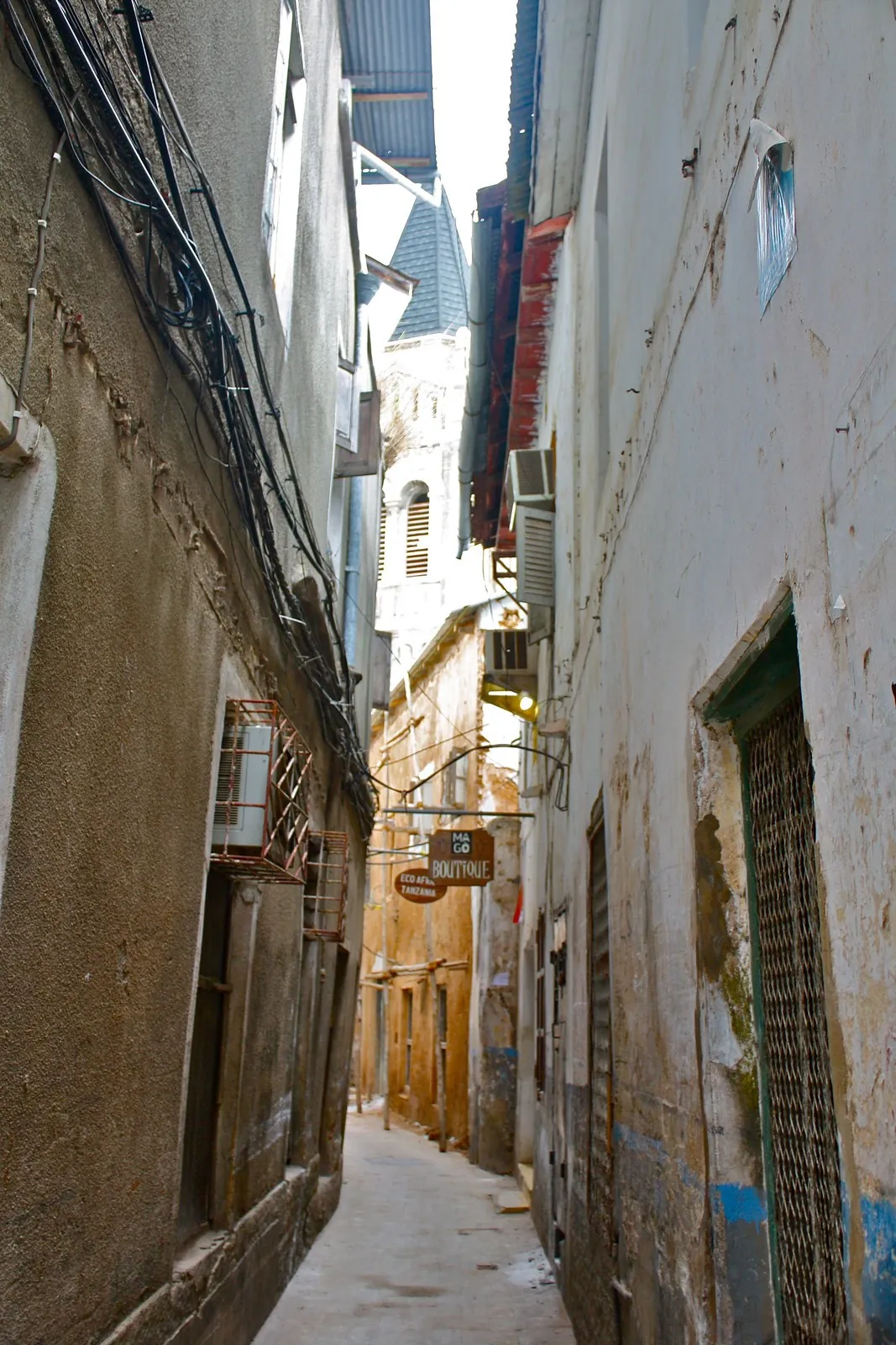 A very narrow alley between high plastered walls with shop signs and a church tower visible above