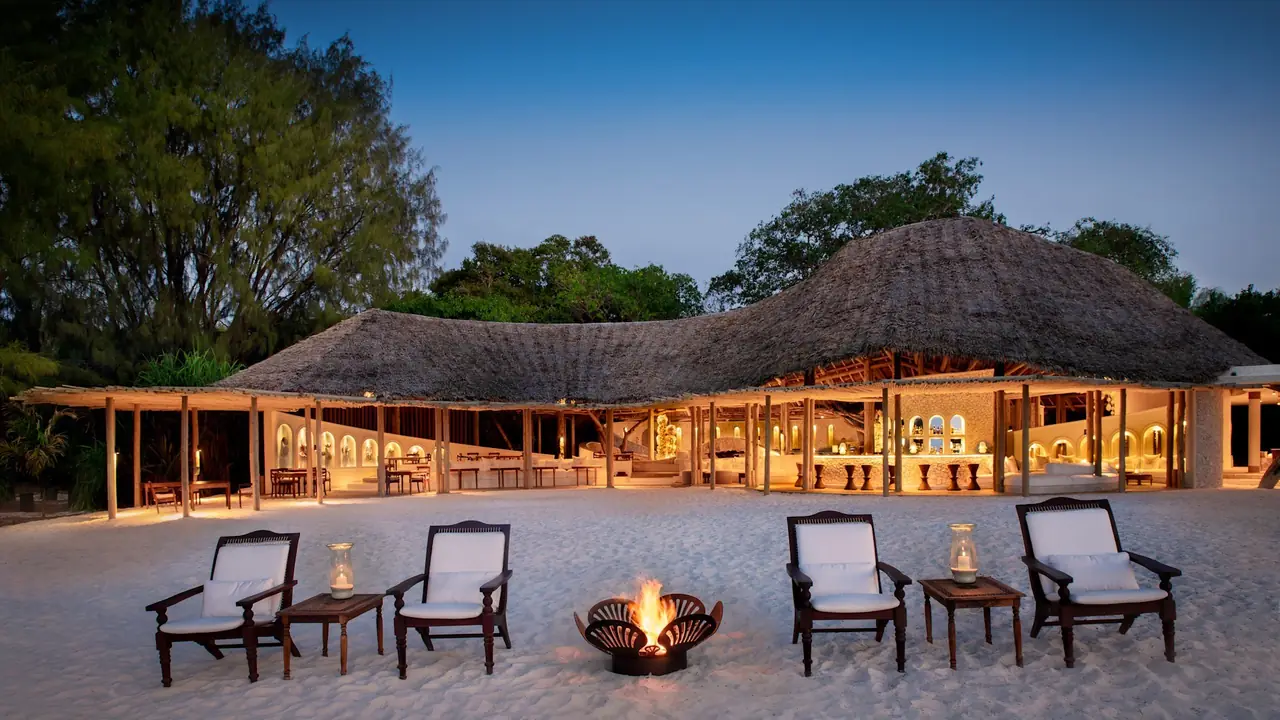 The thatched main lodge lit up at dusk, with chairs around a fire pit on the sand